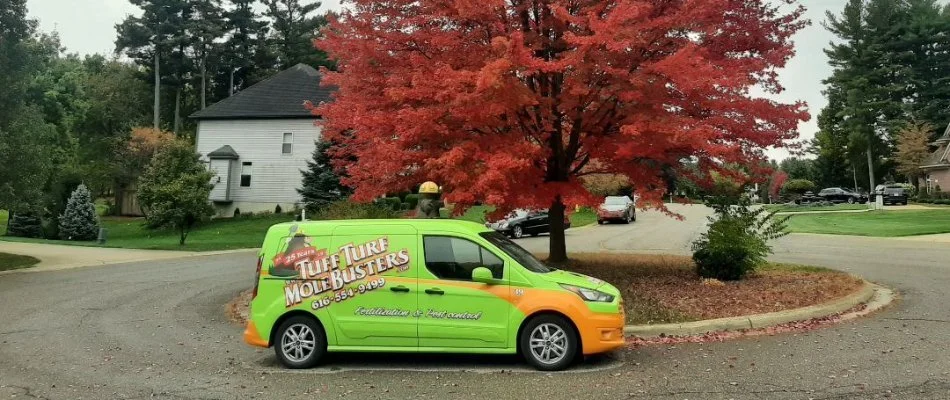 A green and orange 'Tuff Turf Mole Busters' van parked on a road in front of a large house, surrounded by a well-maintained lawn and landscaping