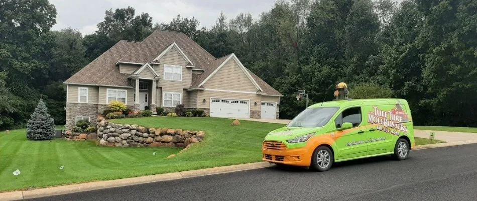 A green and orange 'Tuff Turf Mole Busters' van parked on a road in front of a large house, surrounded by a well-maintained lawn and landscaping